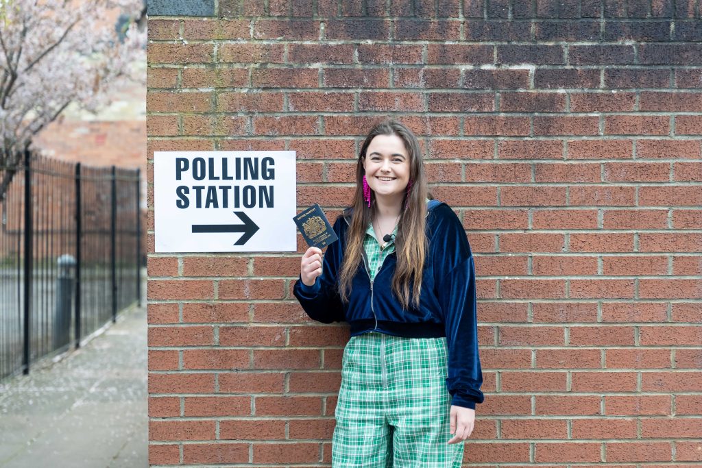 A young voter holds up their passport in front of a sign that says "polling station" with an arrow.
